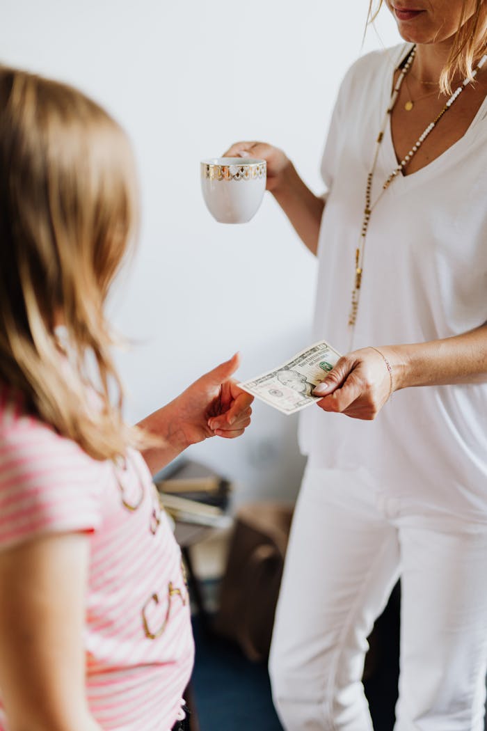 A woman in white hands a dollar bill to a child indoors, symbolizing financial exchange.